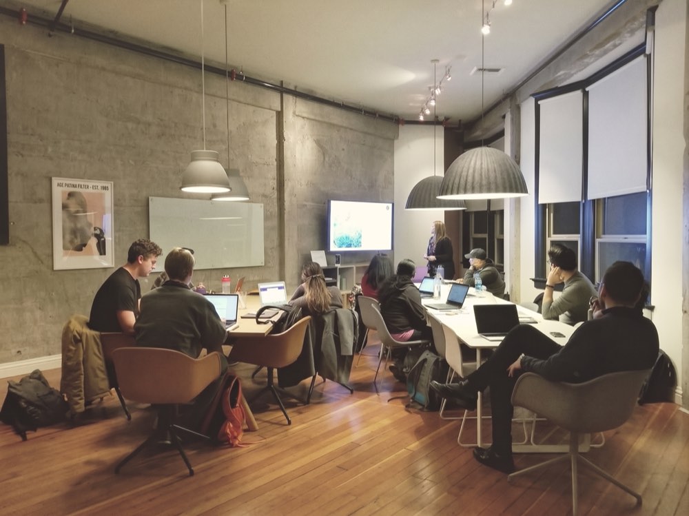Gruppe mit Laptops um einen langen Tisch in einem Loft-Buero, vorn eine Praesentation
